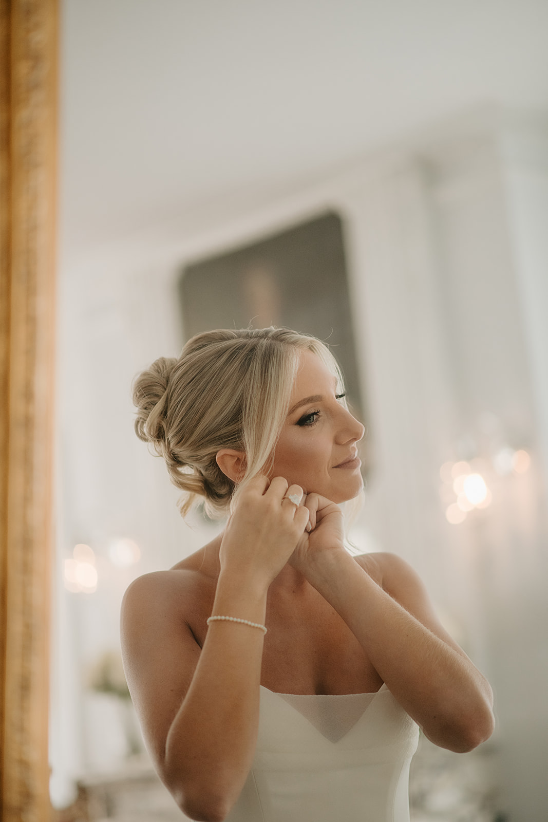 Bride puts on her pearl earrings while looking at her reflection, preparing for her wedding.